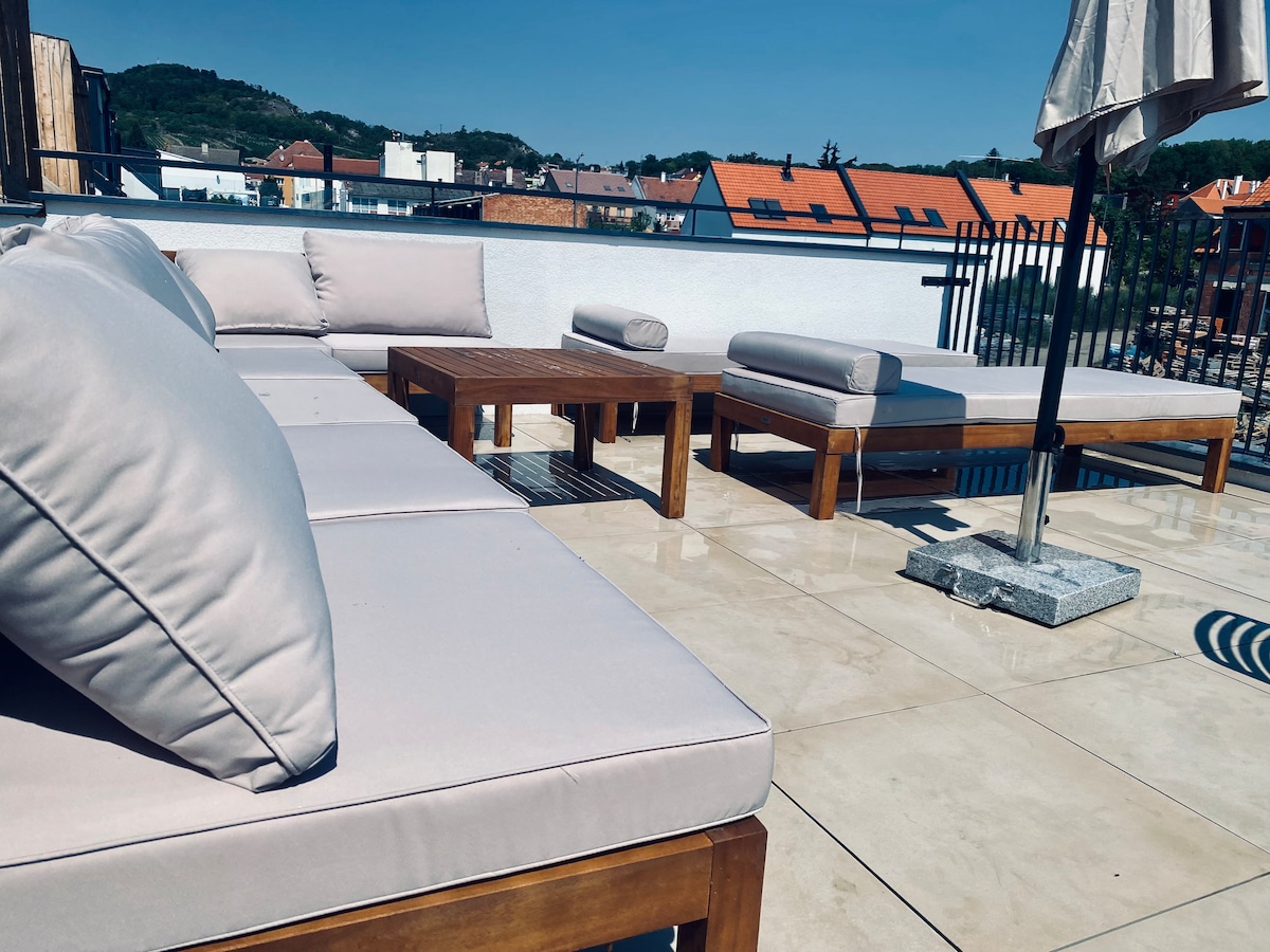 A spacious terrace is furnished with light-colored lounge seating and a coffee table, all set on a tiled surface. An umbrella provides shade, with views of the surrounding landscape visible in the background.