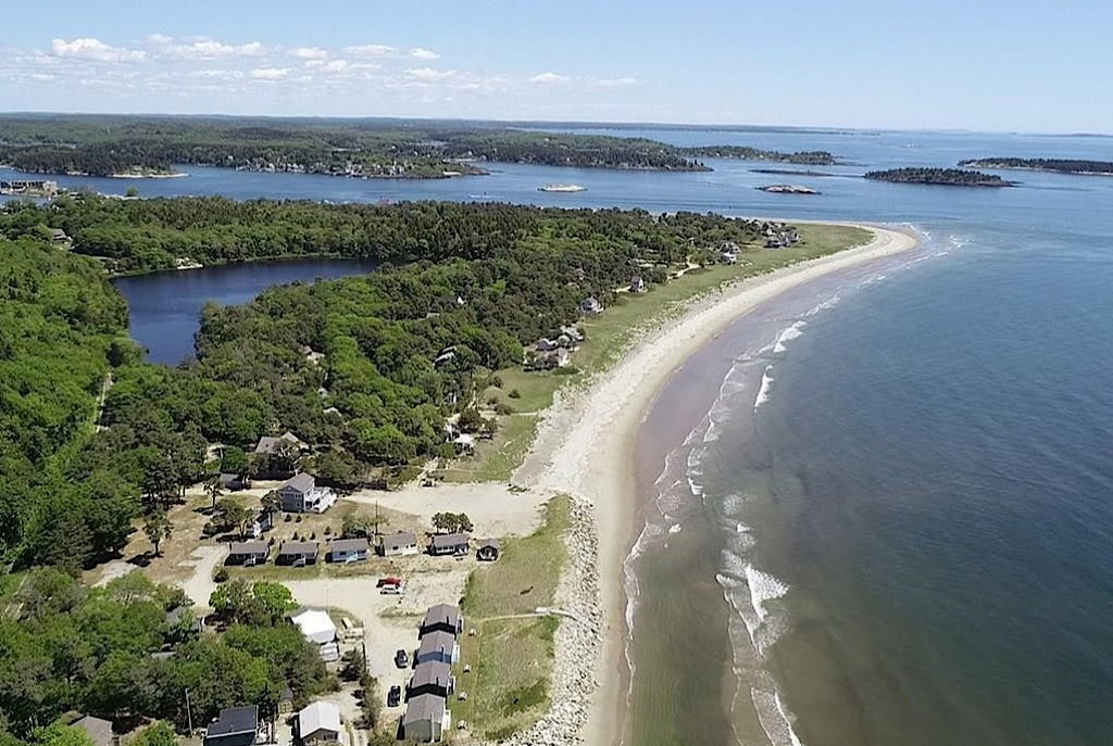 Aerial view of a sandy beach stretching along the coastline, surrounded by lush greenery and water. The image showcases the curve of the shoreline, with various cottages and homes visible near the beach and a serene body of water partially seen behind the trees.