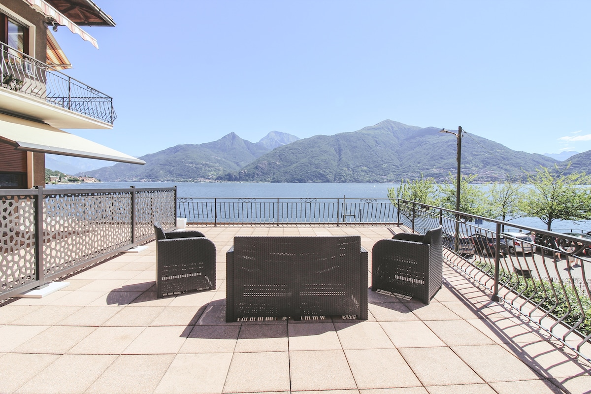 A spacious terrace offers views of the surrounding mountains and lake. Outdoor seating is provided with a set of four wicker chairs arranged around a low table, all positioned on a tiled surface. The railing adds security while maintaining the panoramic sight.