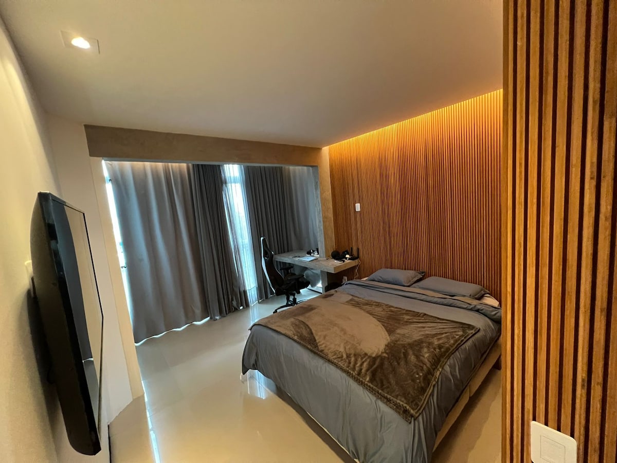 The bedroom features a spacious bed covered with soft bedding. A modern desk and ergonomic chair are positioned beside a large window, allowing for natural light. Textured wooden panels add visual interest to one wall, while a flat-screen TV is mounted opposite the bed.