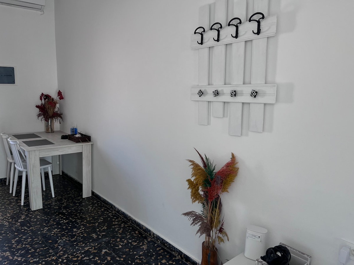 A bright interior space features a minimalist wooden table paired with white chairs. A decorative wall display of six hooks is positioned above a plant, adding color to the area. The dark floor contrasts with the light walls, creating a clean and open atmosphere.