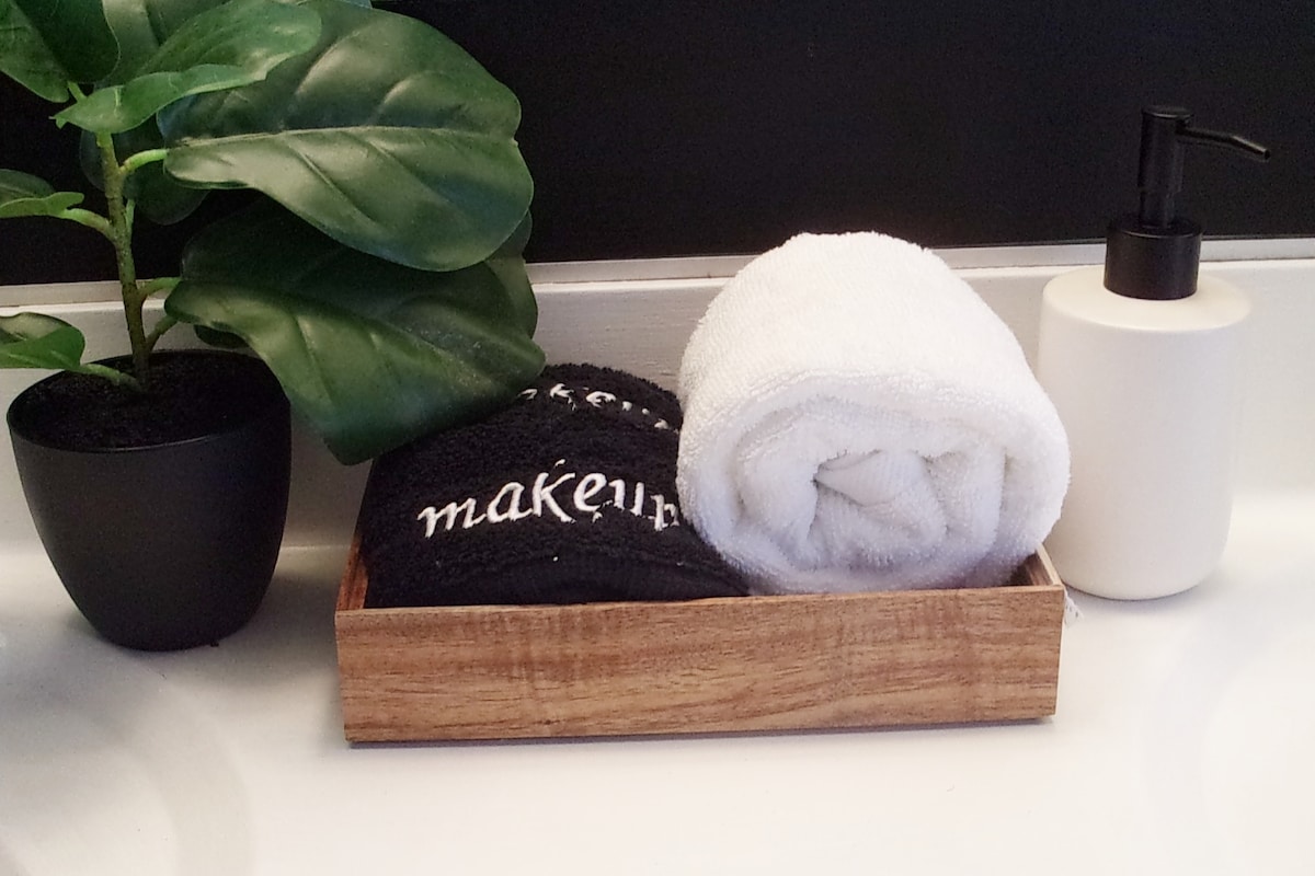 A wooden tray is displayed on a bathroom countertop, holding black and white towels along with a black container labeled 'makeup.' A small potted plant and a white soap dispenser are also present, contributing to a clean and organized appearance.