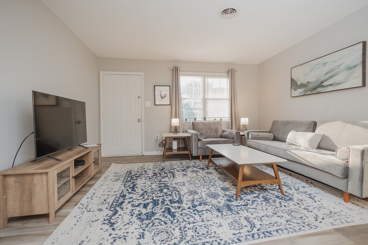 A comfortable living room is presented, featuring a sectional sofa and an armchair arranged around a wooden coffee table. A large flat-screen TV is placed on a media console. Soft natural light enters through the windows, complemented by two table lamps. A patterned area rug adds warmth to the space.