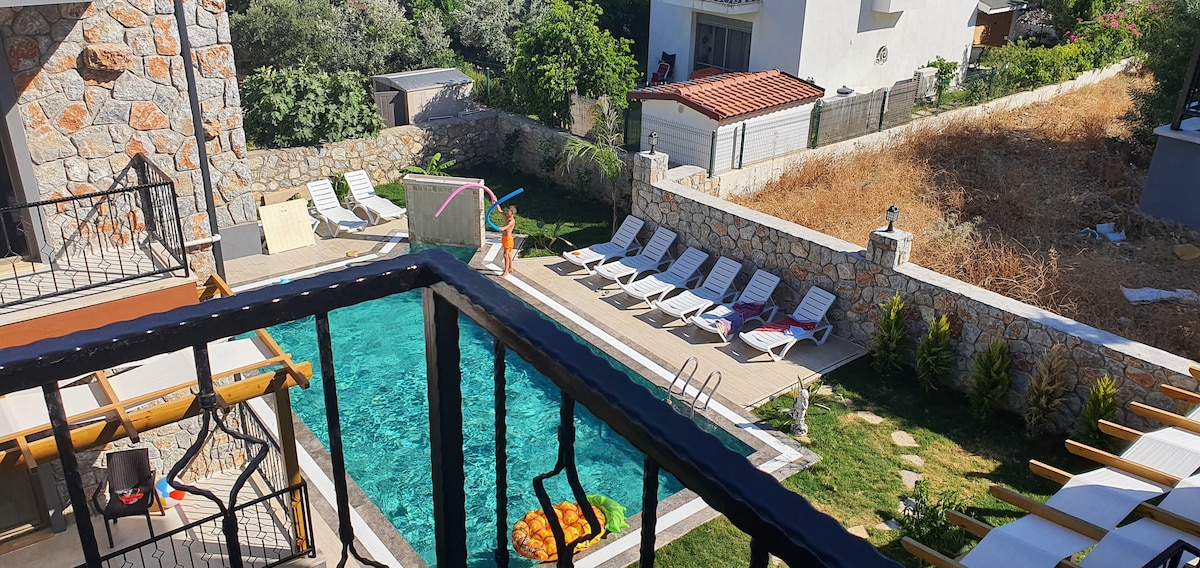 A view from a balcony reveals a private pool surrounded by sun loungers arranged neatly on a deck. Lush greenery and partially landscaped areas are visible beyond the pool, with a structure in the background providing additional space.