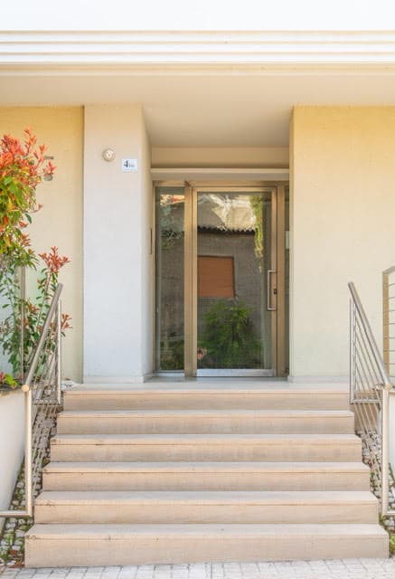 The entrance features a set of stairs leading up to a glass door, framed by a modern facade. Potted plants flanking the steps add a touch of greenery. The surrounding area is well-maintained, contributing to an inviting approach.