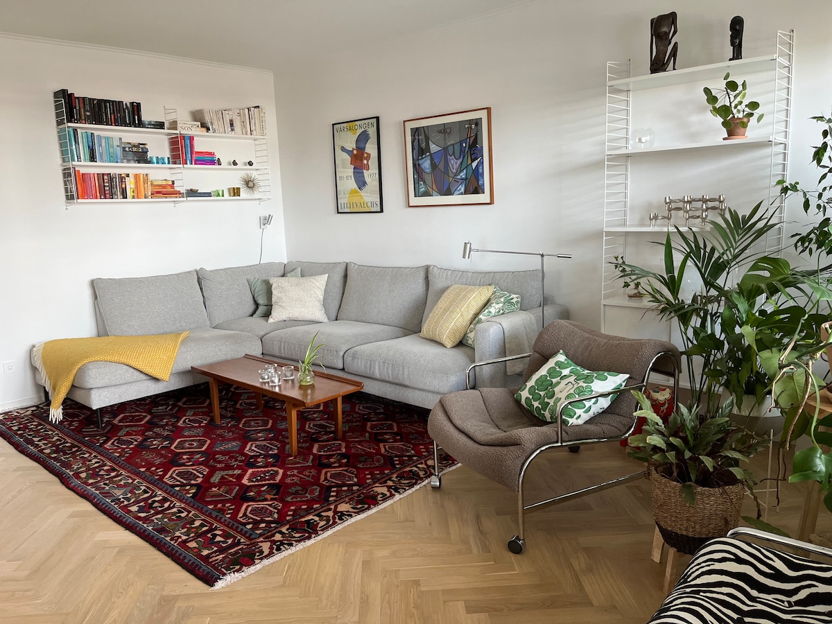 A spacious living area features a large gray sectional sofa paired with a cozy armchair. A wooden coffee table sits in front, surrounded by an area rug. Bookshelves display a diverse collection of books and decor. Plants add a touch of greenery to the room.