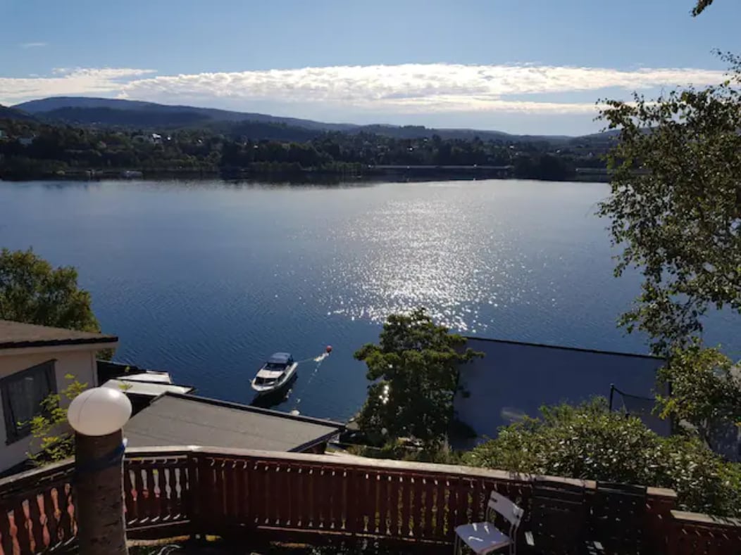 A serene view of a calm fjord is displayed, with sunlight glistening on the water's surface. A small boat is anchored nearby, surrounded by greenery. The foreground features a wooden deck with railing, enhancing the connection to the scenic landscape.