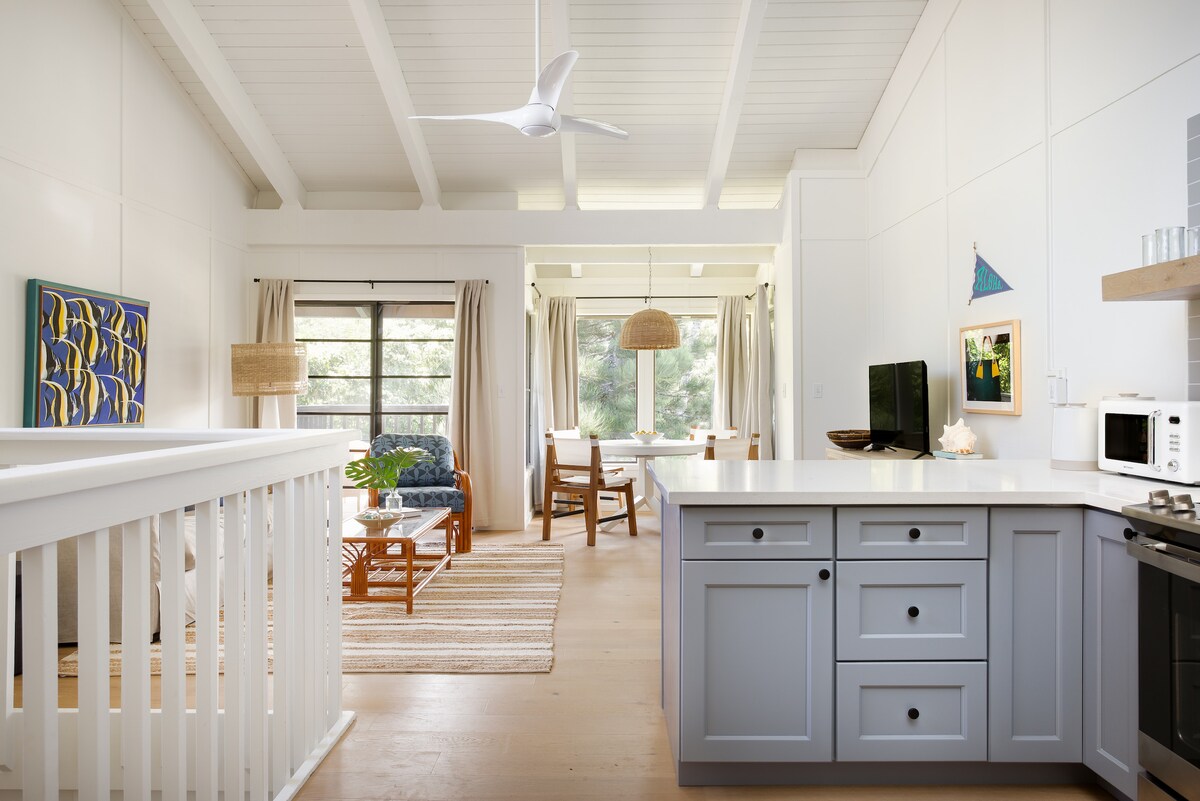 An airy kitchen is highlighted by light grey cabinetry and modern appliances. A ceiling fan creates airflow, while natural light pours in from large windows, illuminating the open living space that includes rattan furniture and decorative wall art.