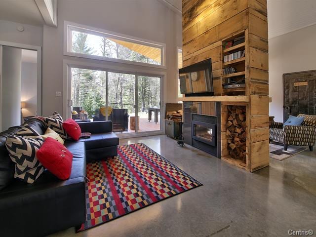 A modern living area features a black leather sofa adorned with colorful cushions, positioned on a patterned rug. A wooden accent wall supports a TV, with firewood stacked below. Large windows offer a view of the outdoor greenery, enhancing the bright and airy ambiance.