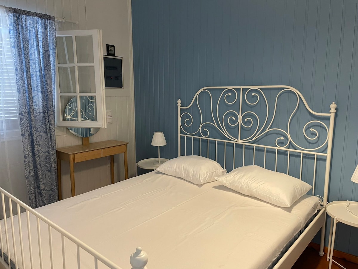 A spacious bed with a white metal frame is positioned against a light blue wall. Two plush white pillows are neatly placed on the bed. A mirror with a decorative frame and a small wooden table are visible beside the bed, accompanied by two bedside lamps.