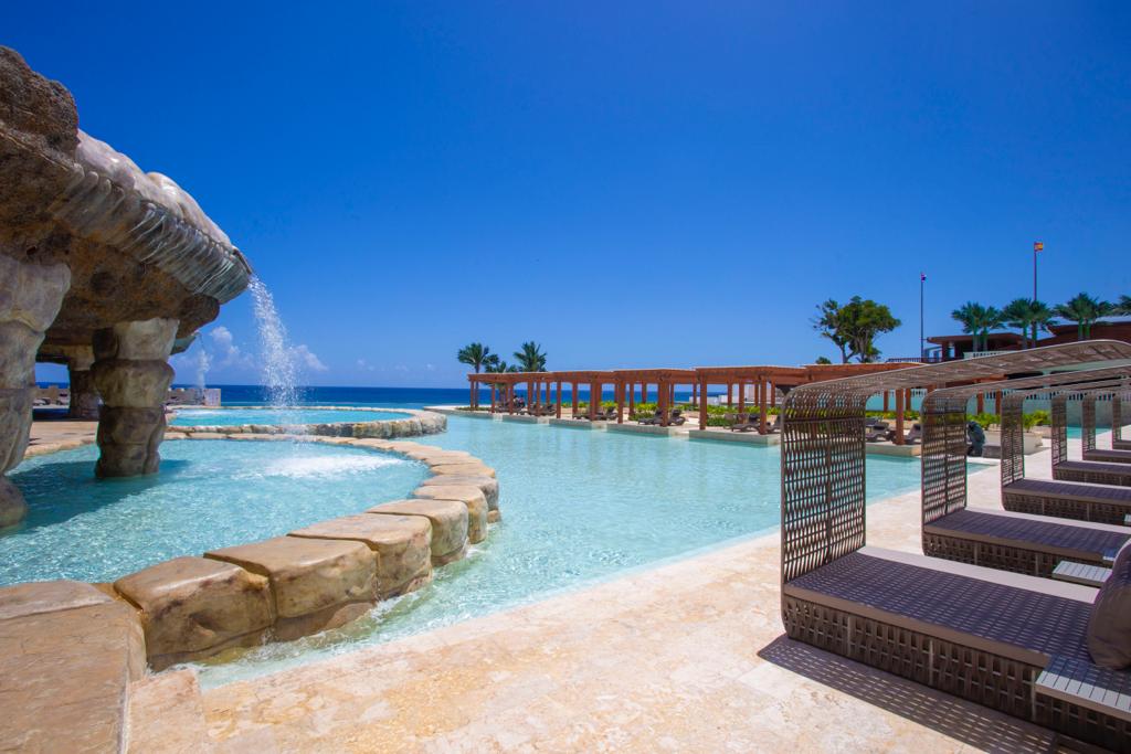 A large outdoor pool area features unique rocky formations and a gentle waterfall, with lounge chairs positioned nearby. A clear blue sky complements the tranquil water, while palm trees are visible along the distant shoreline.