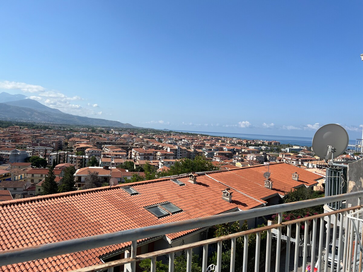 A panoramic view of Monticello showcases terracotta rooftops stretching across the landscape under a clear blue sky. The sea is visible in the distance, with mountains framing the scene to the left, creating a tranquil backdrop.