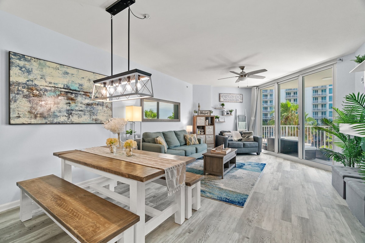 The living area features a spacious layout with modern furnishings, including a comfortable couch and armchairs. A rectangular dining table with benches offers seating for meals, while large windows provide views of the balcony and surrounding greenery. Light wood flooring enhances the fresh ambiance.