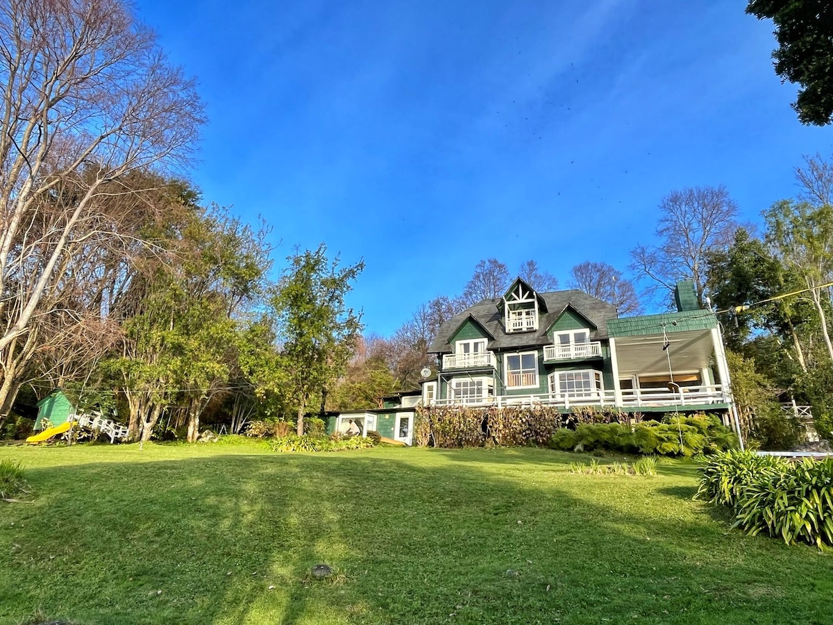 The exterior of the house is surrounded by a spacious lawn, complemented by scattered trees. A large deck extends from the main entrance, and a green canopy is visible in the distance. The well-maintained garden enhances the inviting outdoor area.