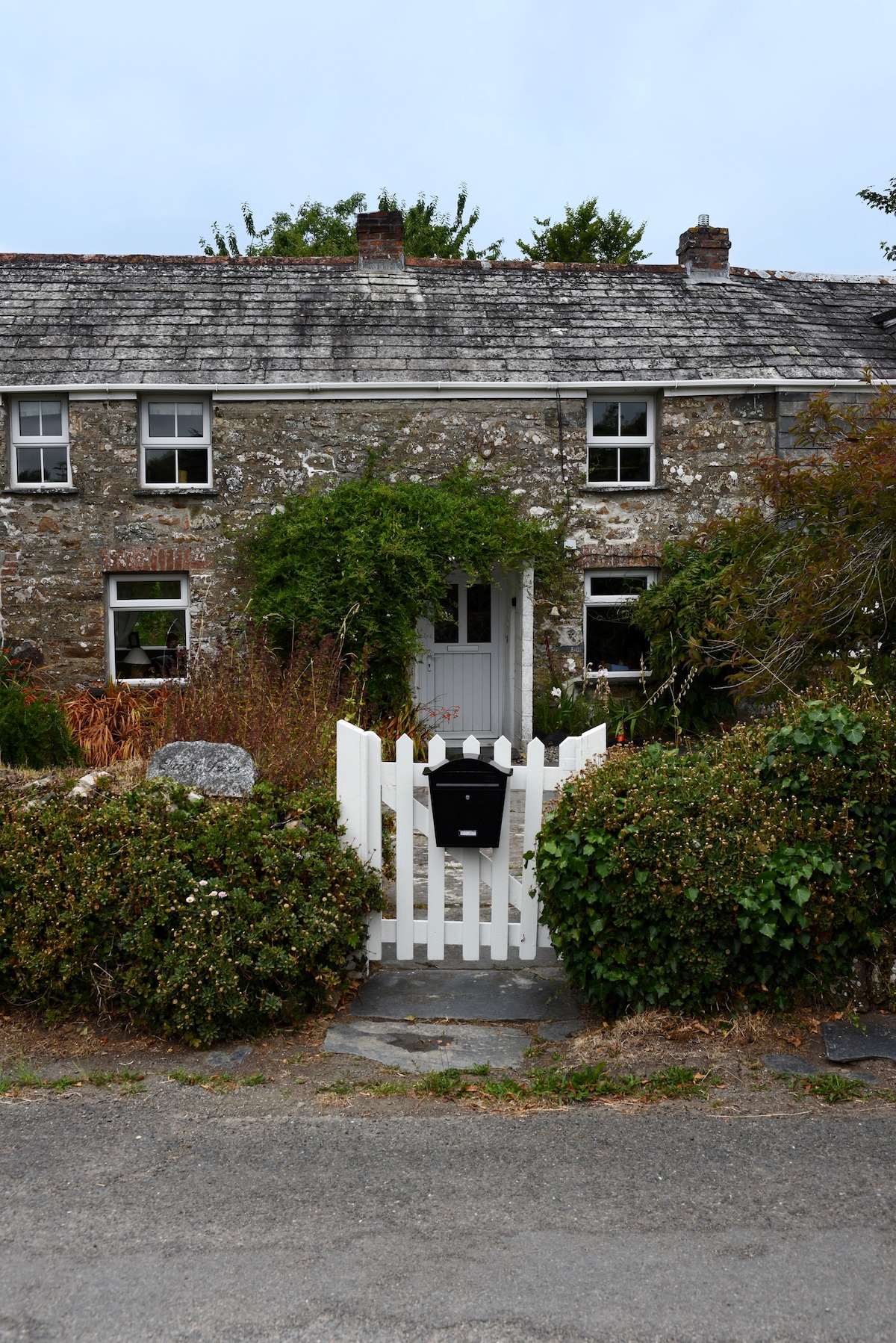 An exterior view of a charming stone cottage features a white picket fence and a gravel pathway leading to the entrance. The house is surrounded by greenery and showcases several windows, with climbing plants decorating the facade.