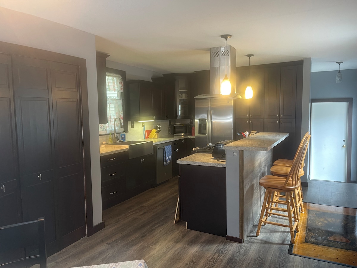 The gourmet kitchen features dark cabinets and a large countertop with seating for three. Stainless steel appliances, including a refrigerator and microwave, are visible, alongside ample cabinet storage. A welcoming atmosphere, created by warm lighting, enhances the functional space.