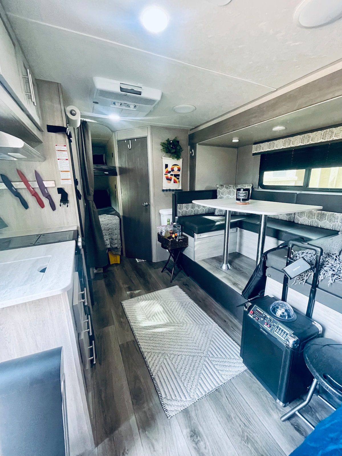 The interior of the camper features a bright, open layout. A dining table with comfortable seating is positioned near the kitchen, which includes modern cooking utensils. The cozy sleeping area is accessible, and a rug enhances the inviting atmosphere of the space.