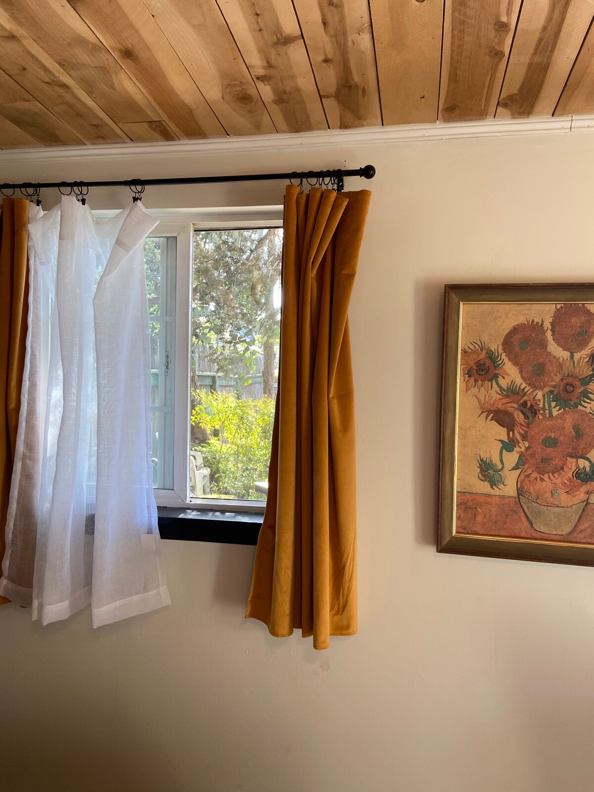 Natural light filters through a window adorned with sheer and vibrant curtains. A floral painting hangs on the wall, adding a touch of color. The wooden ceiling is visible above, enhancing the overall warmth of the space.