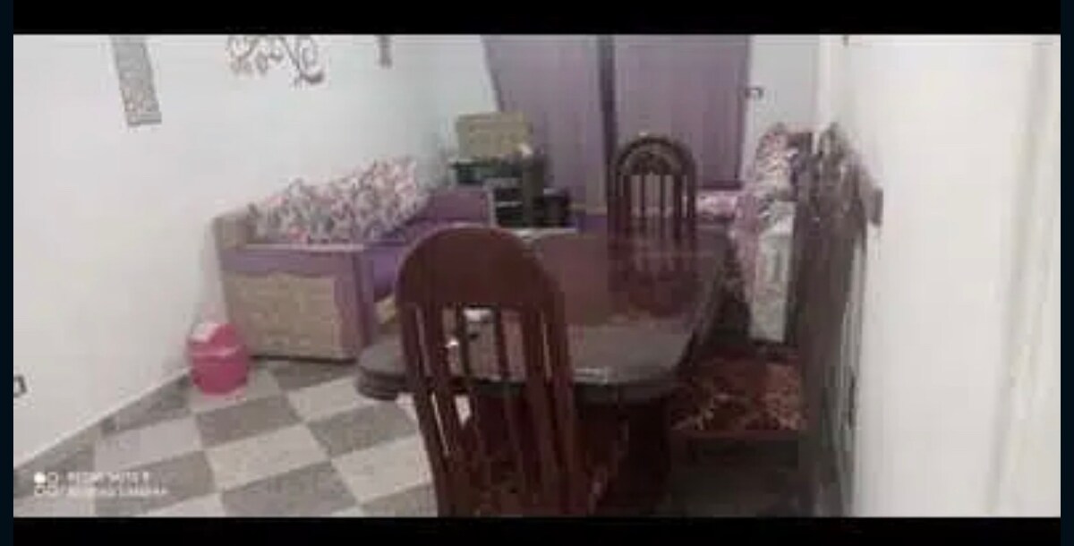 A dining area features a dark wooden table surrounded by several chairs. A plush sofa is visible in the background, complemented by patterned cushions. Light-colored curtains frame the window, while the floor is covered with a checkered tile pattern.
