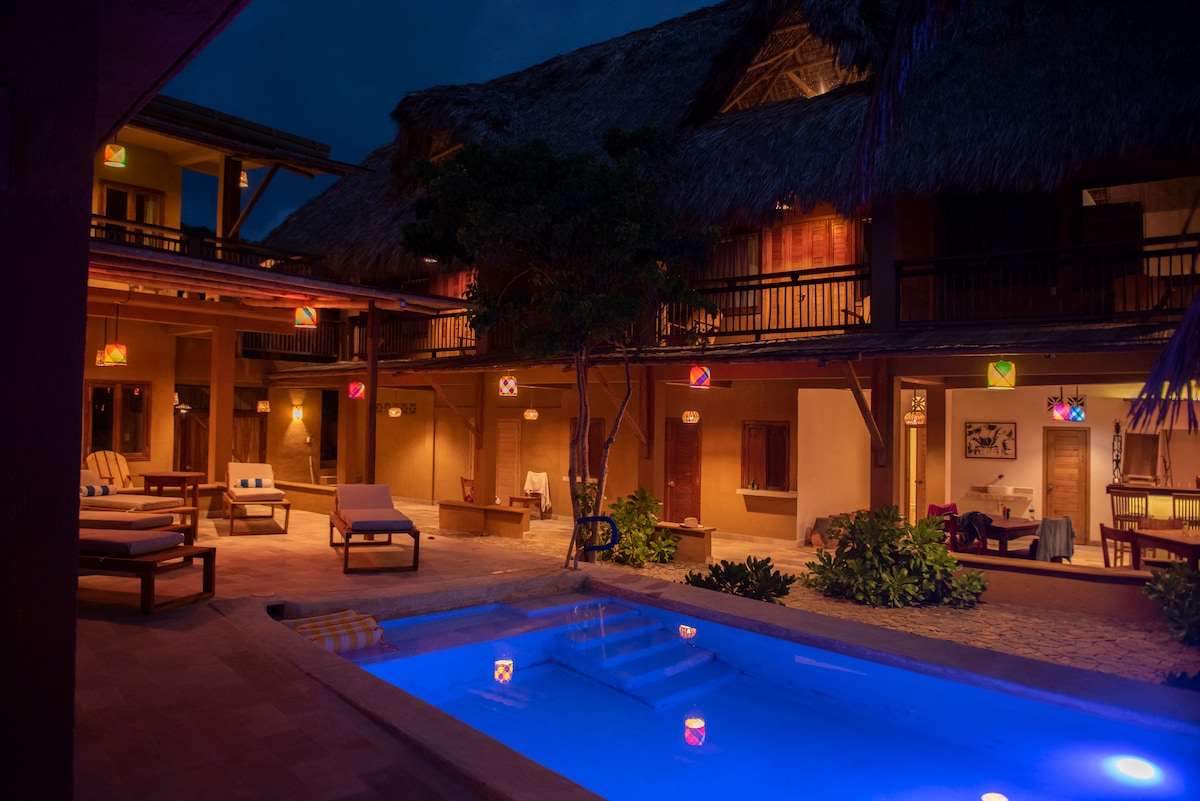 An inviting outdoor space features softly illuminated lounge areas next to a swimming pool. The warm glow from decorative lights complements the rustic wooden architecture and tropical vegetation. The pool area is thoughtfully designed for relaxation within a serene ambiance.