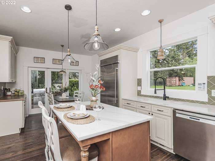 A modern kitchen is featured, showcasing a large island with seating for four. Sleek cabinetry and stainless-steel appliances enhance functionality. Natural light floods the space through a window that overlooks the yard. Decor elements include decorative glasses and floral arrangements on the island.