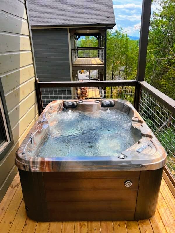 A hot tub is positioned on a wooden balcony, with water gently rippling on its surface. Views of nearby trees and distant mountains are visible, while additional balconies of surrounding cabins can be seen in the background.