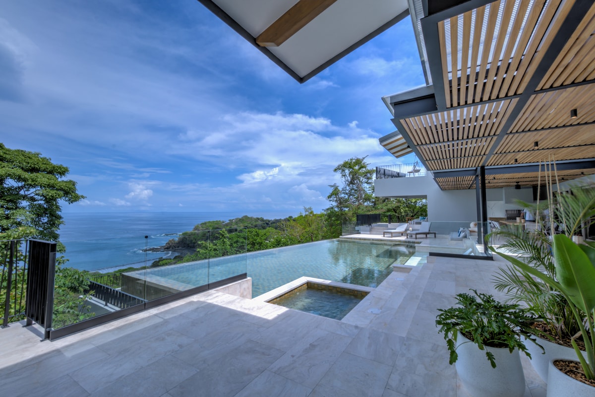 An infinity pool extends toward the edge of the terrace, offering an unobstructed view of the ocean and surrounding greenery. The outdoor area features sleek seating arrangements and a covered space with wooden slats, providing shade while enhancing the connection between indoor and outdoor living.