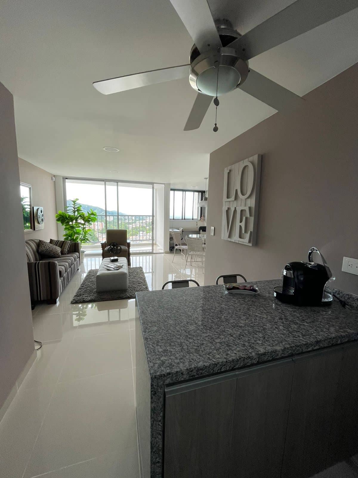 A modern living space features a sleek kitchen island with a granite countertop. A ceiling fan hangs above, providing a cooling breeze. Natural light pours in through expansive glass doors, revealing views of the outdoors. A comfortable seating area is arranged nearby, adorned with a decorative plant.