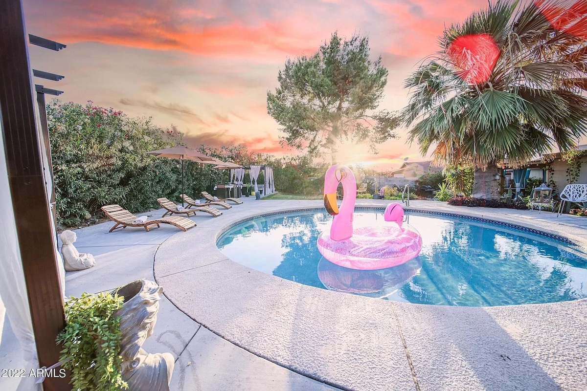 A private backyard showcases a sparkling pool surrounded by lounge chairs and lush greenery. Two vibrant flamingo floaties rest on the water's surface. The scene is illuminated by a warm sunset, highlighting the tranquil ambiance and inviting outdoor space.