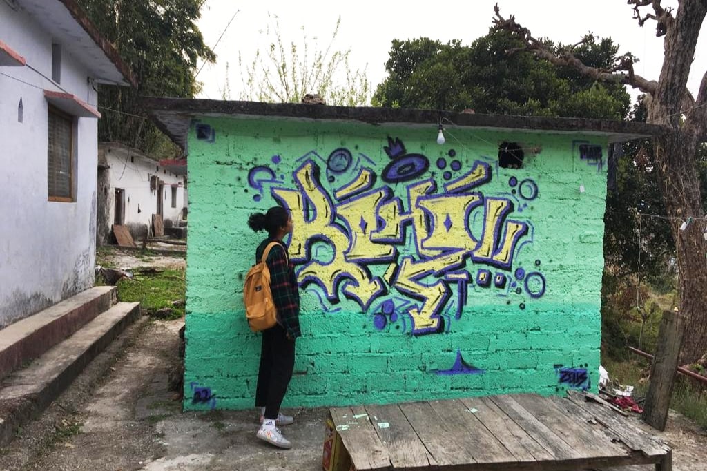 A vibrant mural in shades of purple and blue decorates a green wall. A person stands beside the artwork, facing the wall. In the foreground, a wooden table sits on a gravel surface, with a grassy area and trees visible in the background.