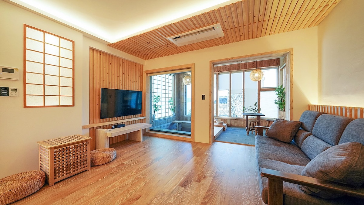 A spacious lounge area features wooden accents and minimalistic decor. A large television is mounted on the wall, opposite a comfortable sofa. Natural light floods in through sliding doors leading to a serene outdoor space, enhancing the peaceful environment.