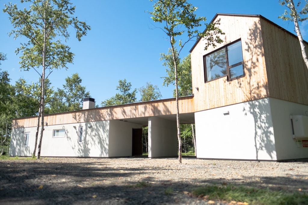 A modern vacation house features a combination of wood and white plaster exterior. Set among trees, the structure has large windows that allow natural light to fill the rooms, while a spacious covered entry area provides shelter from the elements.