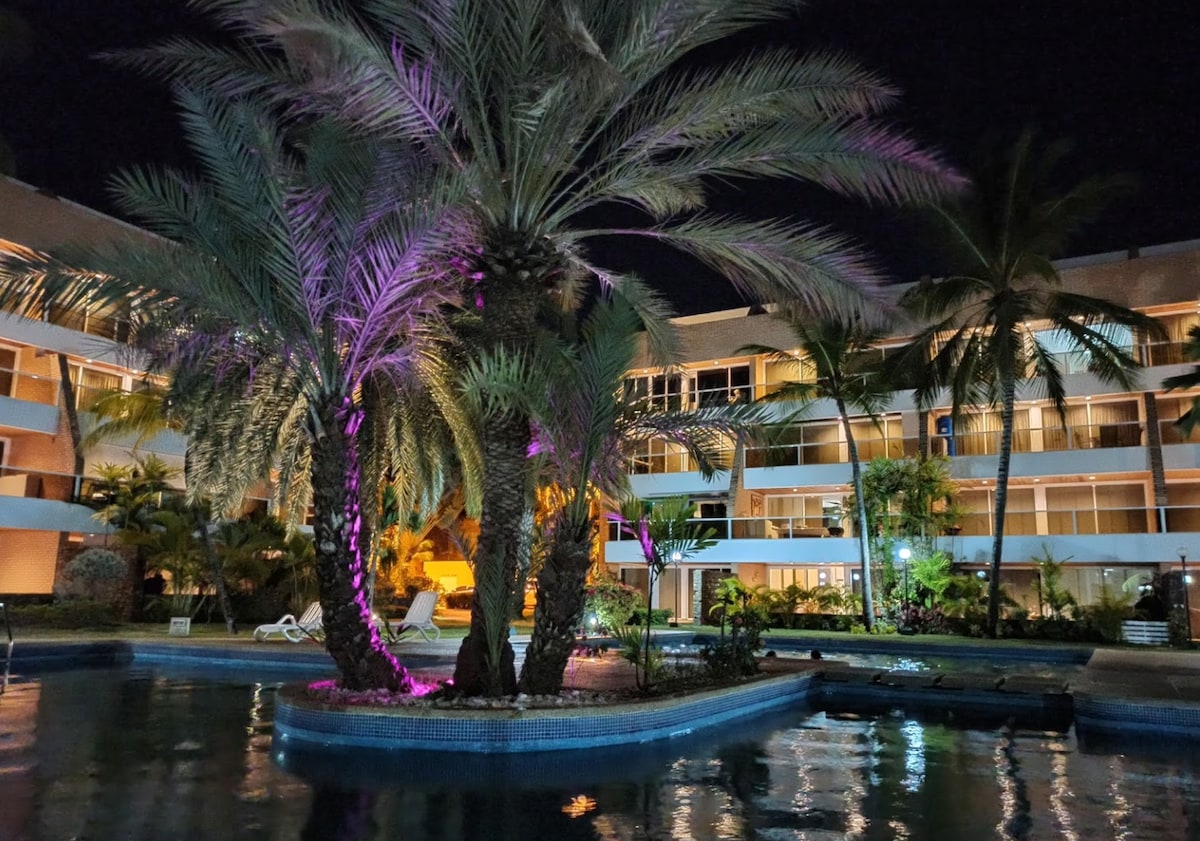 The pool area is surrounded by palm trees illuminated in soft purple hues, creating a serene atmosphere. The multi-level building is seen in the background, featuring balconies with glass railings. Lush greenery complements the tranquil setting, inviting guests to unwind.
