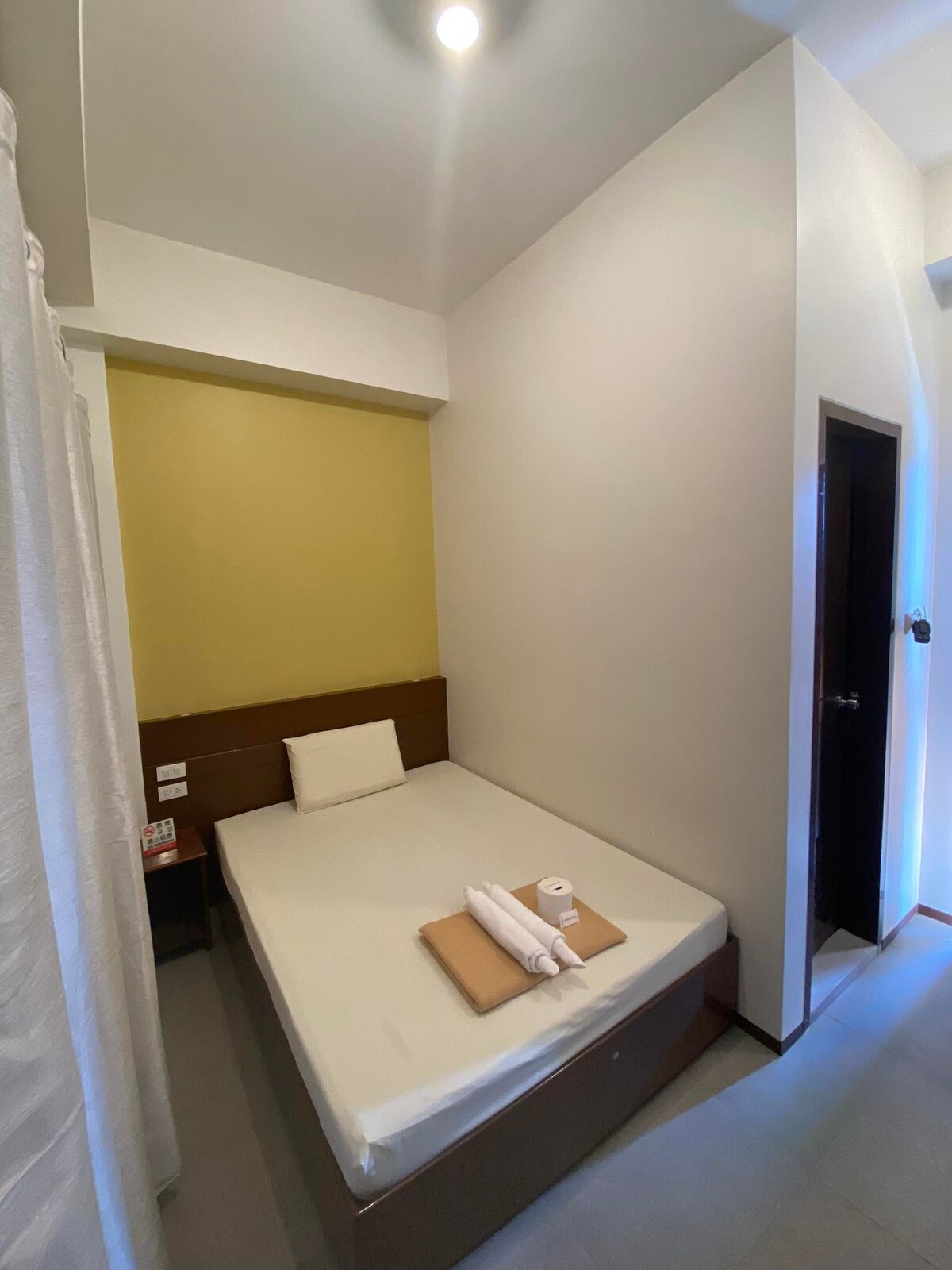 A compact sleeping area features a bed with a neutral-colored bedspread. A cushioned headboard is located against a soft yellow accent wall. A small wooden table holds rolled towels, while a door leading to the private bathroom is visible on the right side.