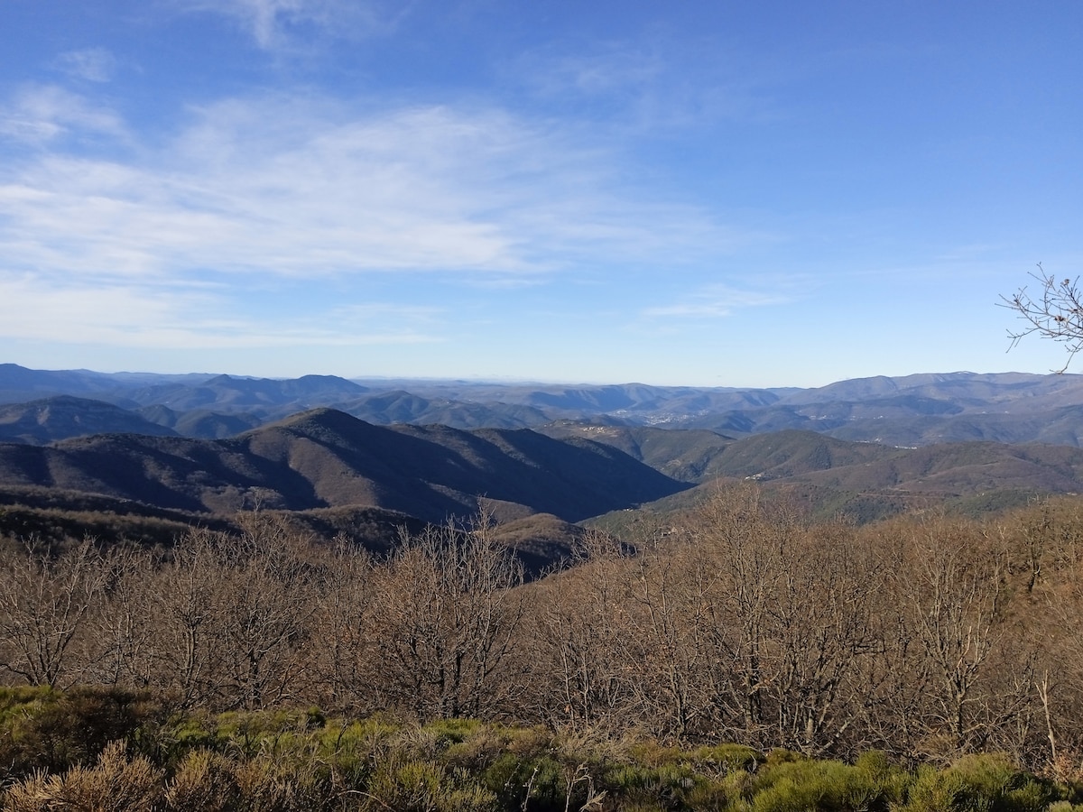 A panoramic view of rolling mountains stretches across the landscape, showcasing varying shades of brown and green foliage. The sky above is clear with soft clouds, and the terrain presents a serene, natural setting that highlights the beauty of the surrounding area.