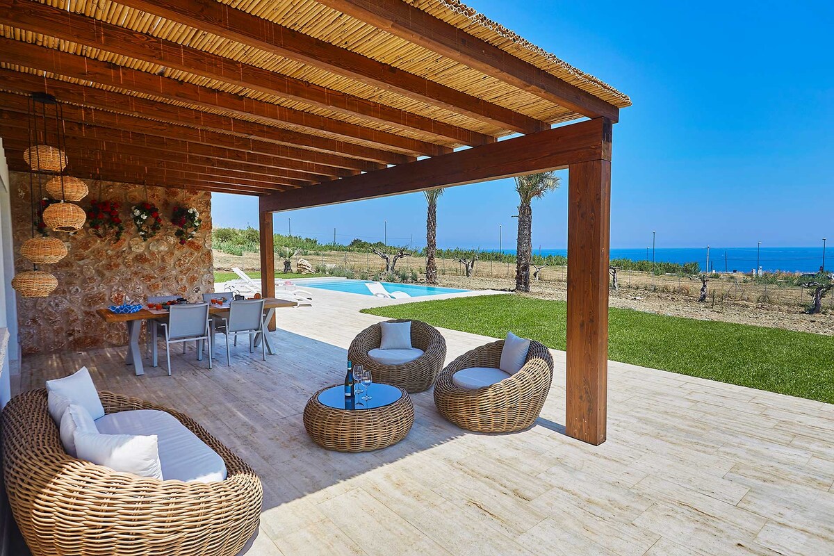 A covered outdoor veranda features modern wicker seating and a dining table, overlooking a serene landscape. The space is bathed in natural light, with views of the distant sea and an inviting infinity pool in the background.