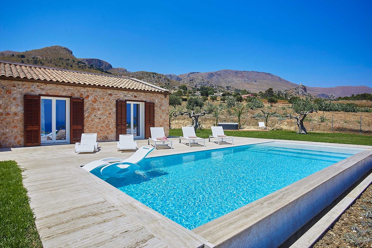 A modern villa is set against a scenic backdrop, featuring a clear, inviting swimming pool with a sleek edge. Sun loungers are arranged around the pool area, which is surrounded by lush green grass and olive trees, creating a serene outdoor retreat.