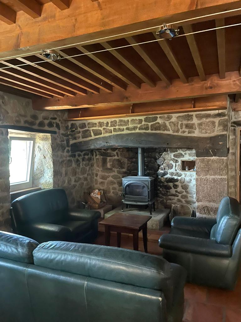 The living space features a cozy seating arrangement with two dark green couches facing a wooden coffee table. A wood-burning stove is situated against a stone wall, providing warmth. Natural light enters through a window, enhancing the rustic charm of the space.