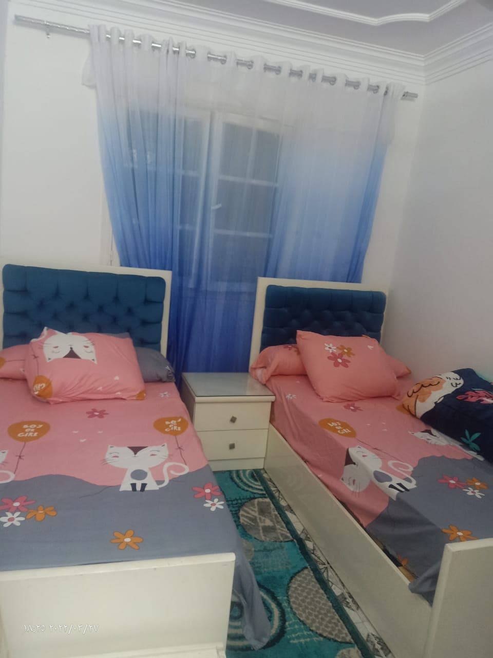 A bright sleeping area features two neatly made twin beds, each adorned with playful animal-themed bedding. A bedside table is positioned between the beds. Light blue curtains frame a nearby window, allowing soft natural light to illuminate the space.