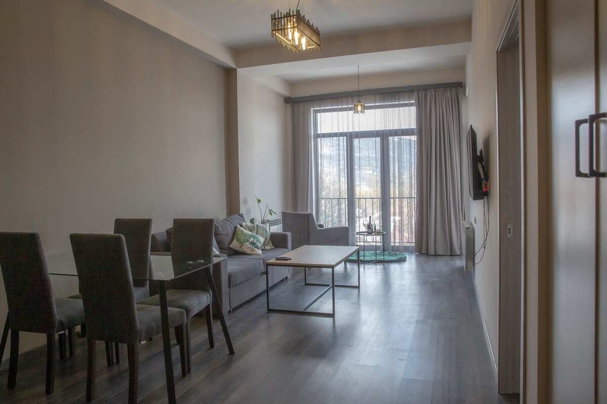 A spacious living room is presented with modern furnishings, including a gray sectional sofa and a glass dining table surrounded by four chairs. Large windows fill the space with natural light, providing access to a balcony with outdoor seating.