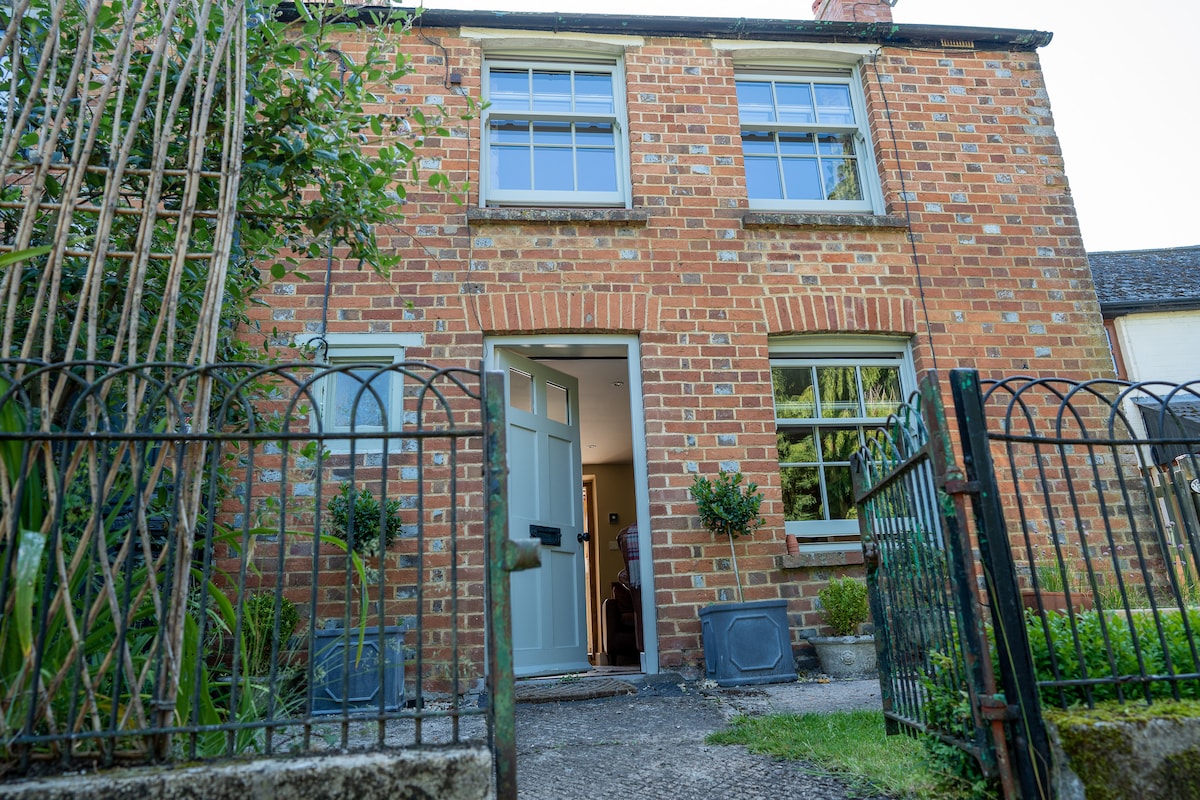 The exterior of the cottage features a charming red brick façade with white window frames. A path leads to the front door, which is painted in a soft blue. Lush greenery and small shrubs are visible beside the path, contributing to the welcoming entrance.
