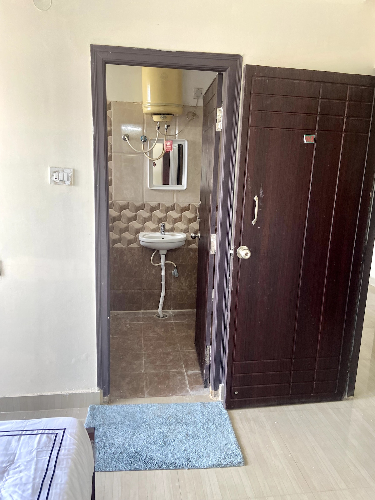 A well-maintained bathroom is visible through an open door, featuring beige wall tiles and modern fixtures. A small mirror is mounted above a basin, and a washing machine connection is located on the wall. The entrance is framed by a dark wooden door and a blue bath mat is placed on the floor.