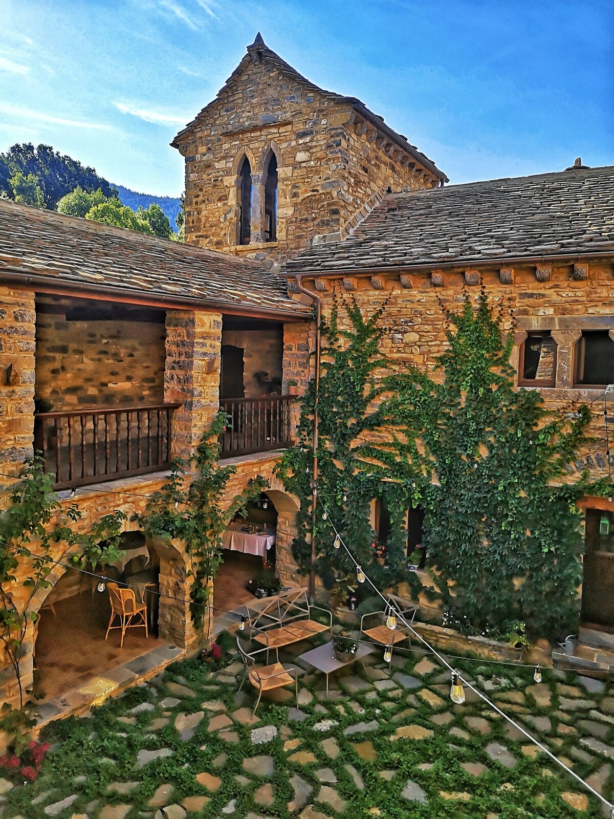 An outdoor courtyard is surrounded by rustic stone buildings characterized by arched doorways and balconies. Green ivy climbs the walls, adding a natural element. A few tables and chairs are arranged on the cobblestone ground, creating an inviting space for relaxation.