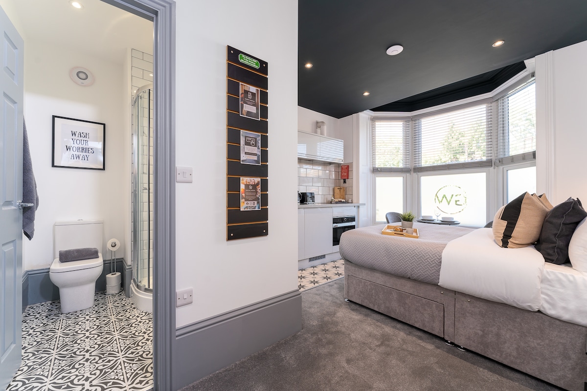 The modern studio interior includes a spacious bed with decorative pillows and a sleek gray blanket. A stylish kitchenette is partially visible, alongside an en-suite bathroom featuring a shower. Large windows bring in natural light, enhancing the overall design of the room.