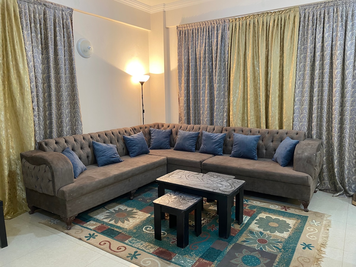 A spacious living area is featured, showcasing a large tufted sectional sofa adorned with blue cushions. A patterned rug lies beneath a set of three black coffee tables. Floor-to-ceiling curtains in gold and silver frame the windows, creating a cozy ambiance.