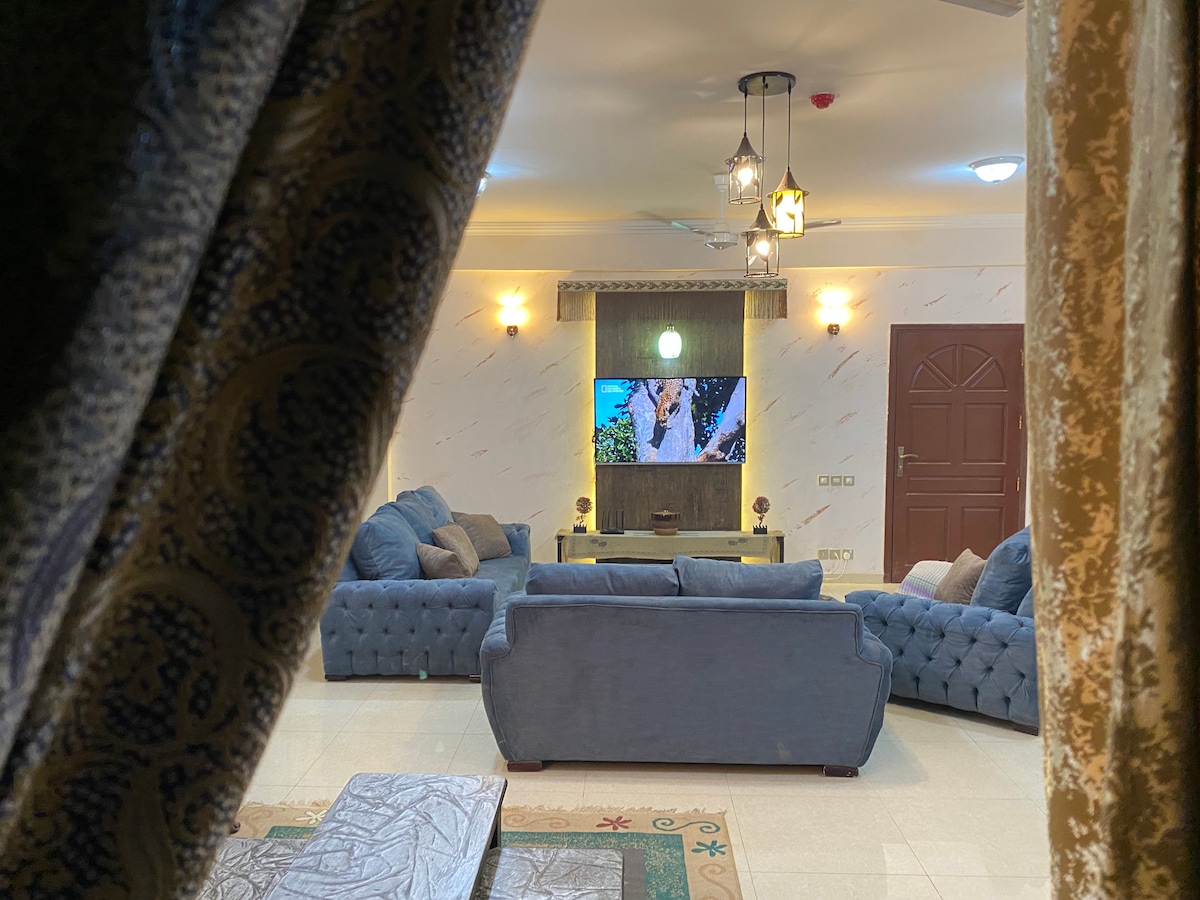 A spacious living area is visible through partly drawn curtains, featuring two large sofas and a coffee table. A television is mounted on the wall, showcasing a program. Soft lighting is provided by multiple fixtures, creating a relaxed environment.