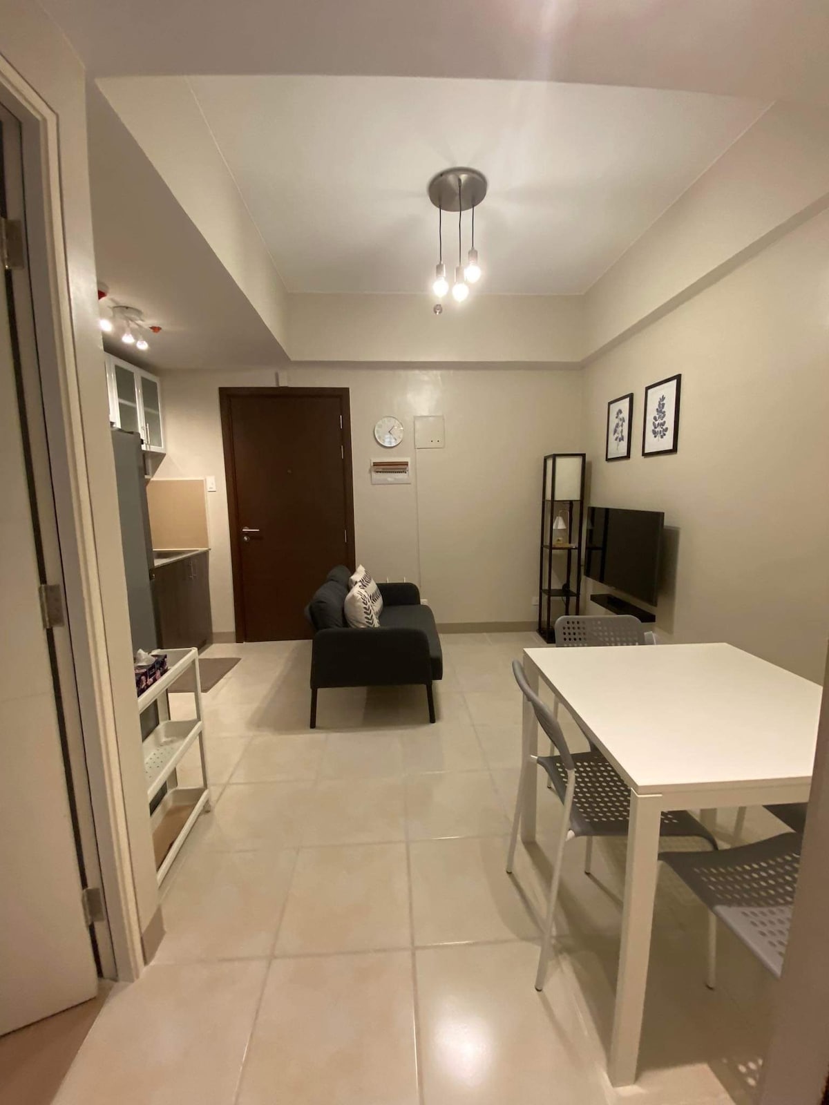 A modern living area includes a dark upholstered sofa facing a wall-mounted Smart TV. A dining table with four chairs is positioned nearby, and a minimalist shelf stands against the wall. Natural light is provided through overhead lighting and an open space.