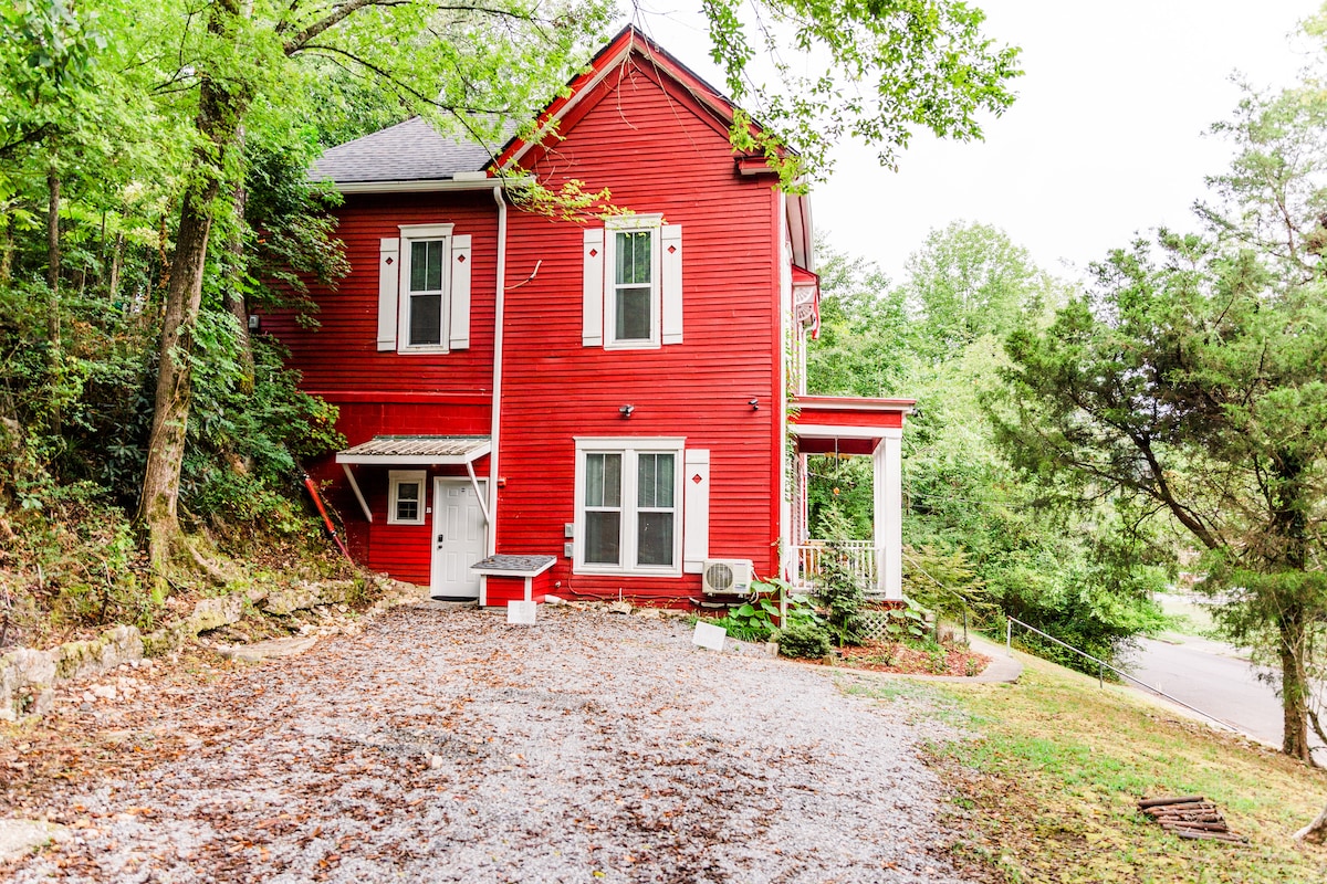 The exterior of a red Craftsman home is set against a backdrop of lush greenery. The building features multiple windows and a welcoming porch, surrounded by a gravel area that offers ample space for outdoor activities. A small air conditioning unit is visible on the side.