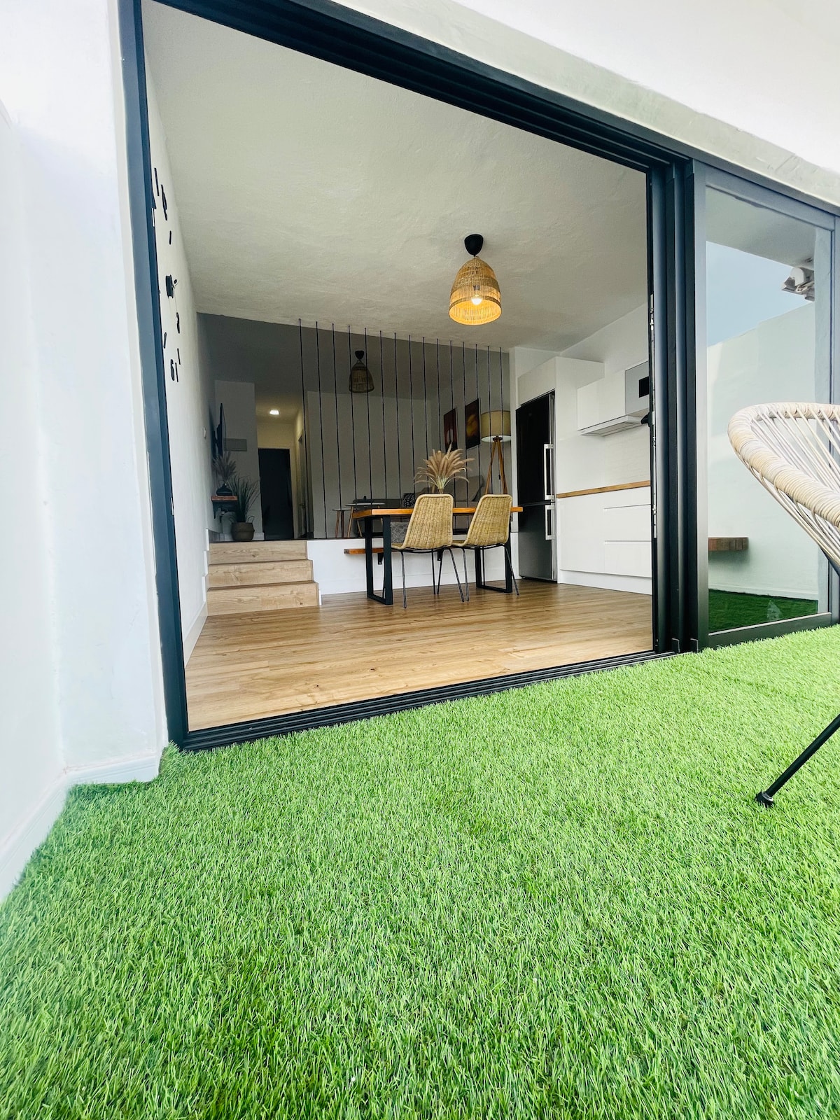 A view from an outdoor space reveals an open dining area featuring a wooden table surrounded by four chairs. The area is complemented by a green artificial grass surface and an overhead light fixture adds warmth. The interior leading to the kitchen is visible through large glass doors.