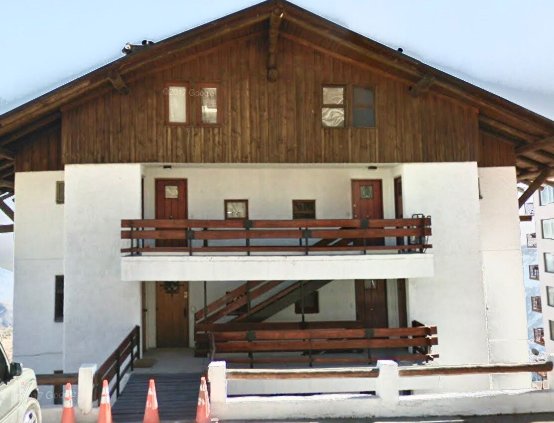 A two-story mountain apartment building features a wooden upper level and a white lower level. Balconies with wooden railings are visible, along with several doors leading into the units. Parking cones are arranged neatly in front of the structure.